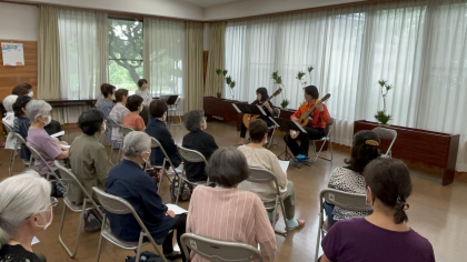 写真：20人ぐらいのご年配の方々が、椅子に座ってギターの生演奏を聴いている様子