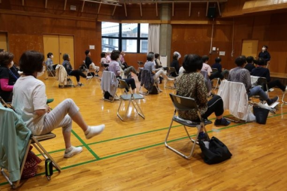 写真：体育館で、ご年配の方々が椅子に座って足上げ運動をされている様子