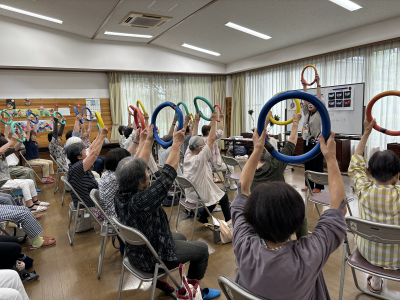 写真：20人ぐらいのご年配の方々が、輪投げの輪っかのようなものを頭上に掲げている様子
